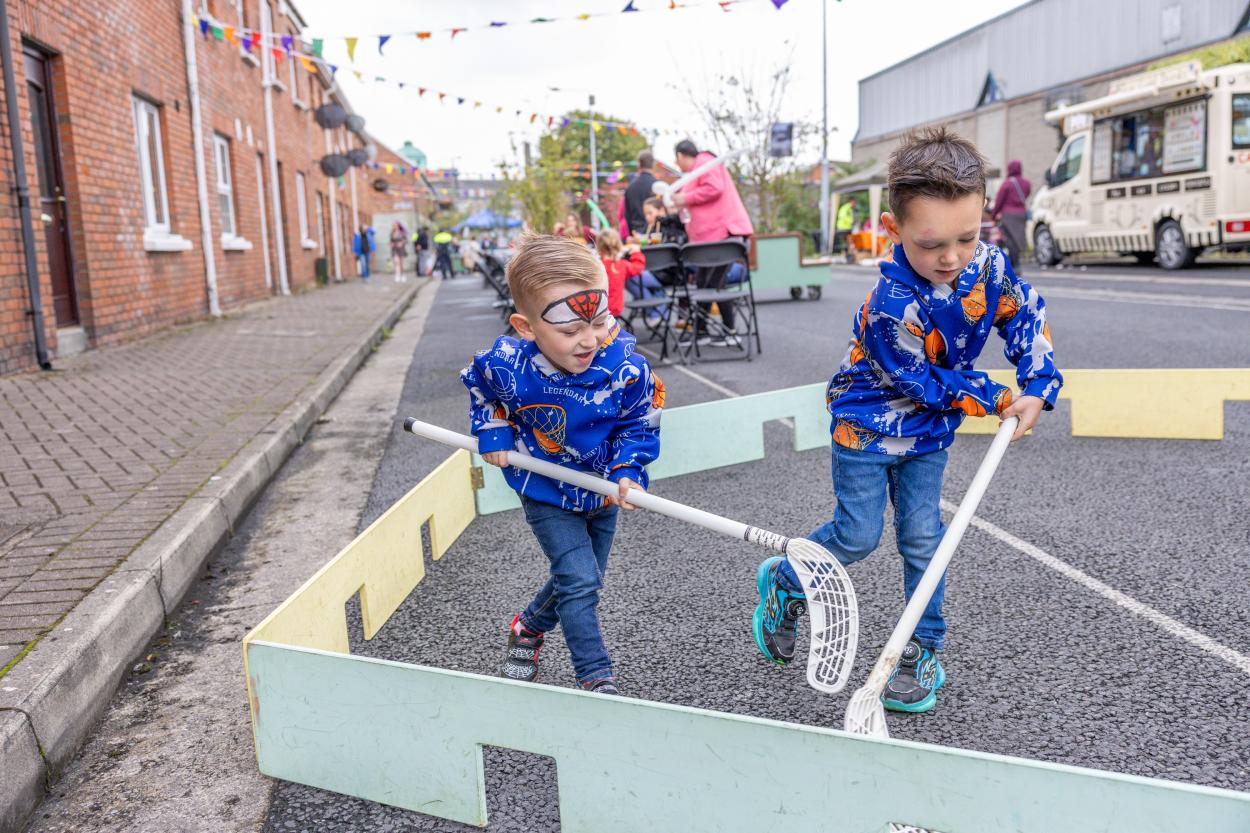 Two young boys in matching blue hoodies play street hockey in a small rink during a lively outdoor festival.