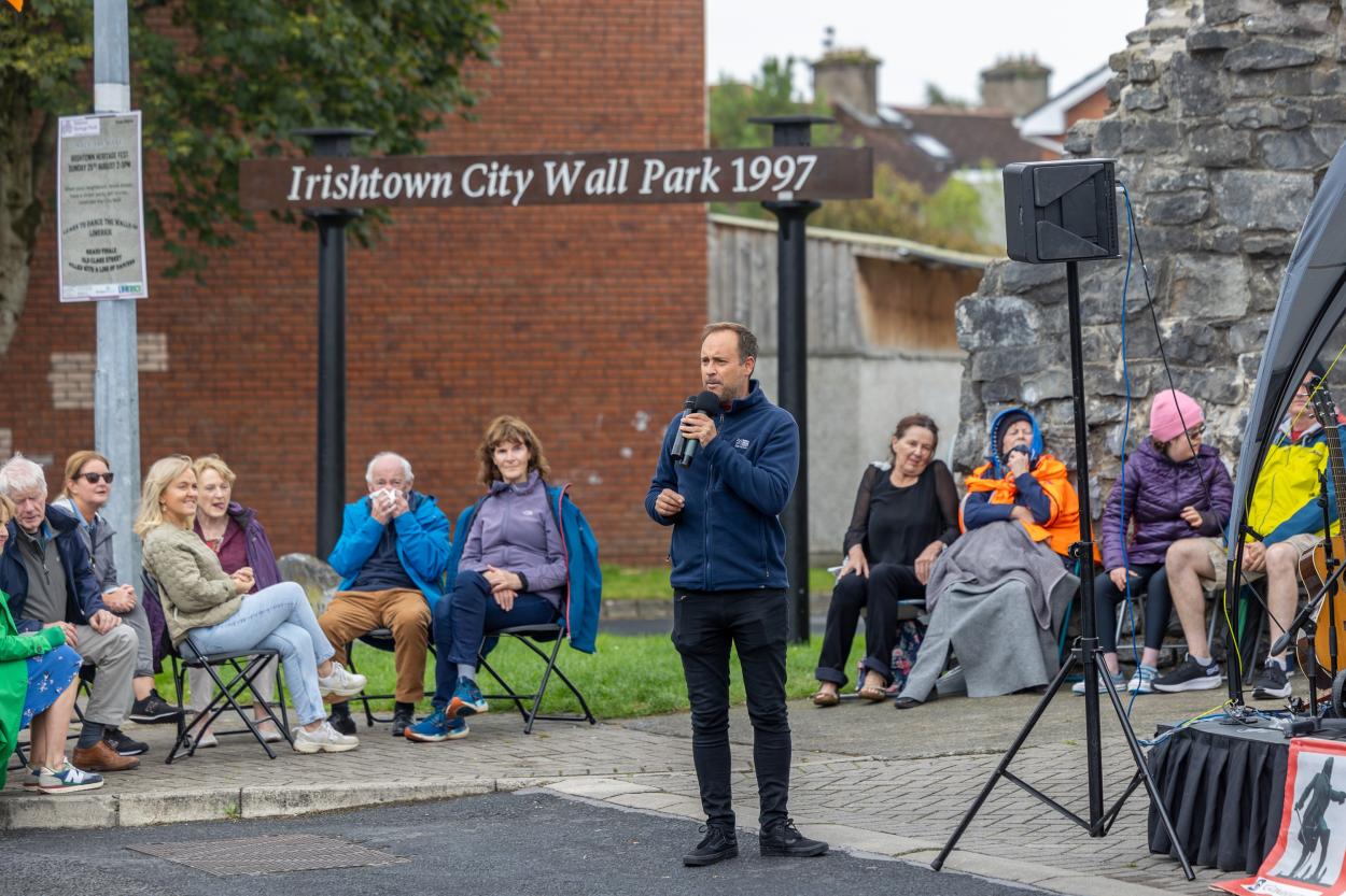 A man speaks into a microphone while people sit on chairs near a stone wall, with a sign reading "Irishtown City Wall Park 1997."