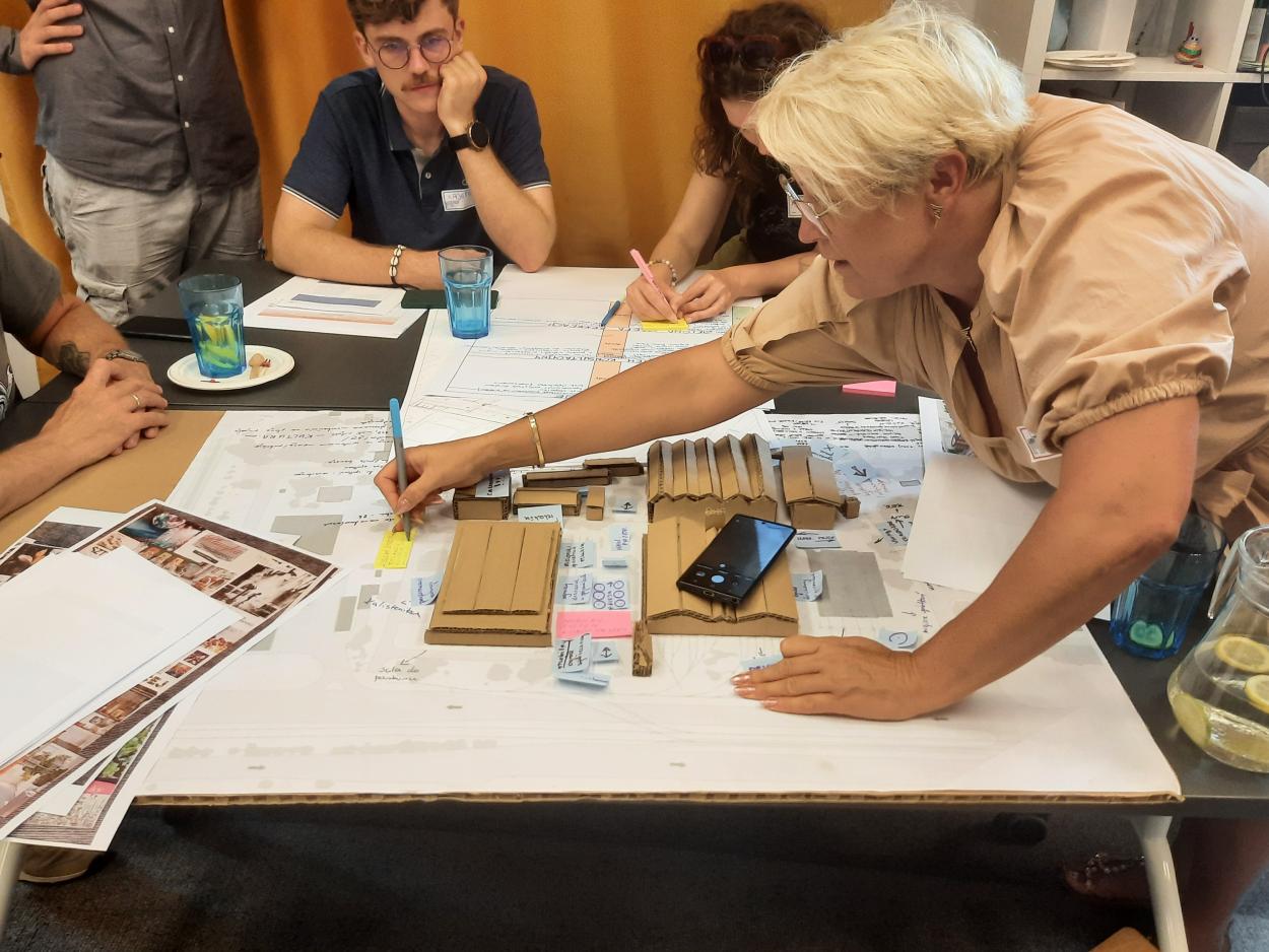 A group of people making notes while sitting/standing around the table with the cardboard depot model in the center.