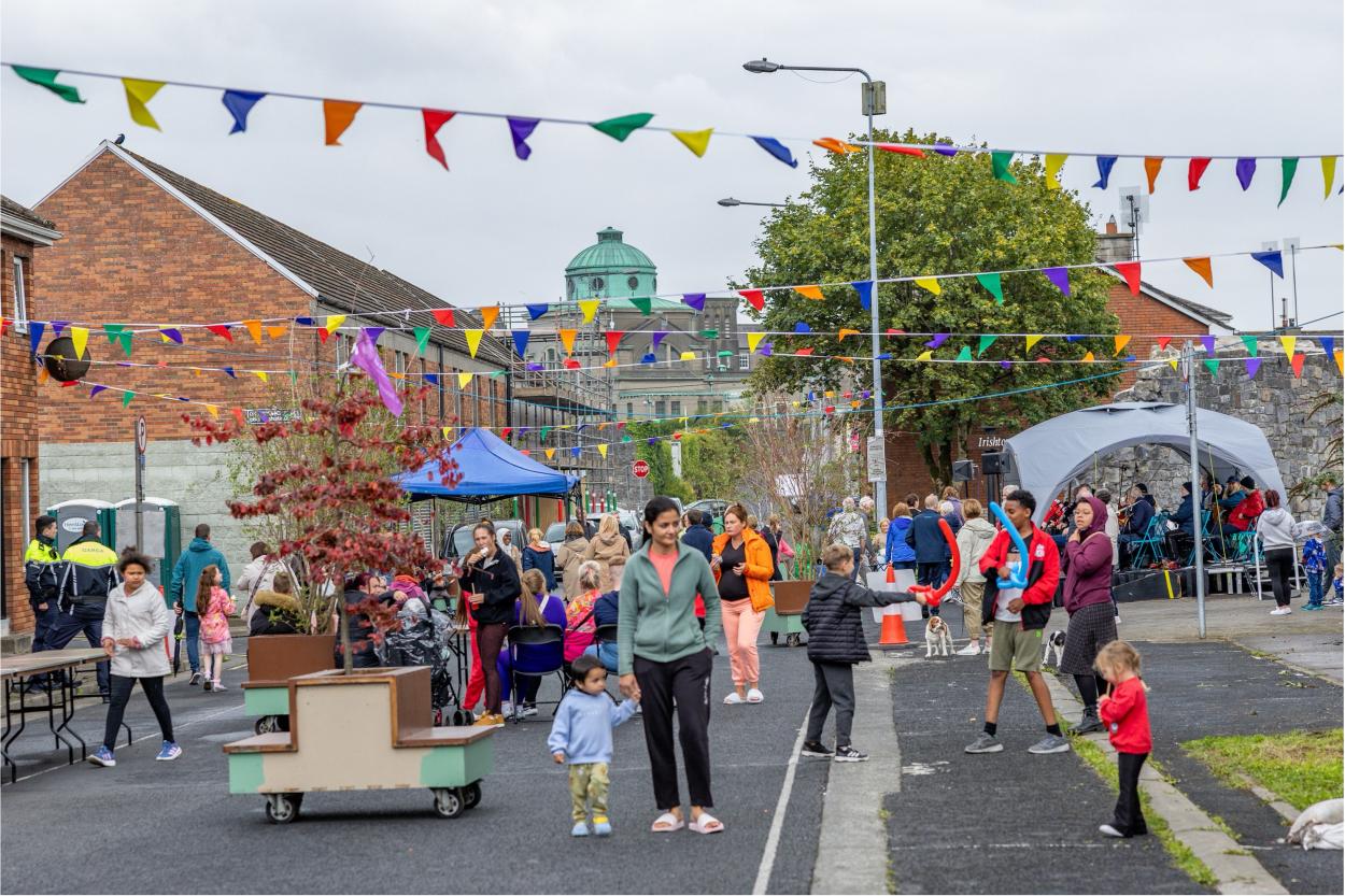 People walking through a street where the festival is held. In the background there are festival tents, mobile planters with benches and colorful pennants hung across the street.