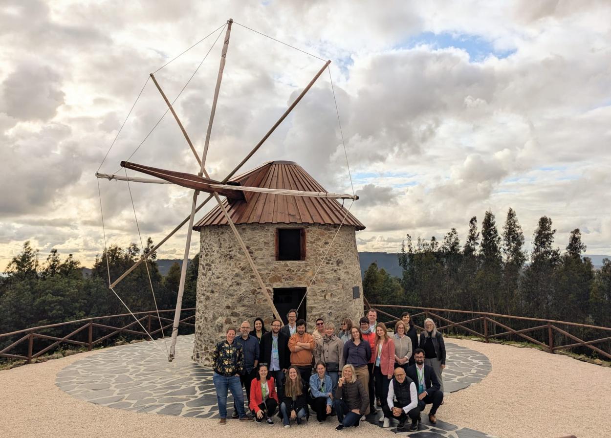 A group of people poses for a photo in front of a stone windmill with wooden sails, set against a mountainous landscape and a cloudy sky. 