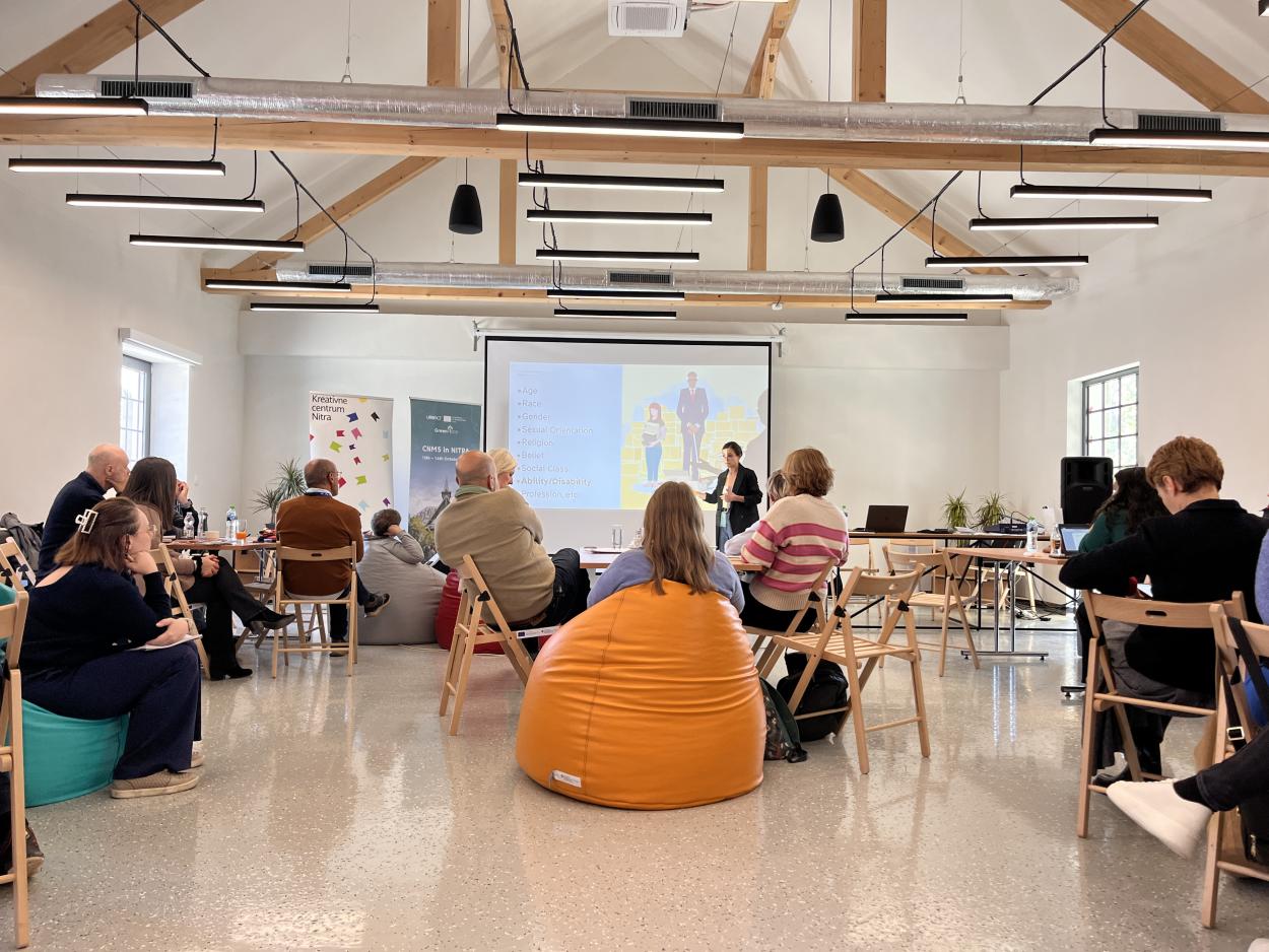 The image shows the participants sitting around tables and listening to the presentation.