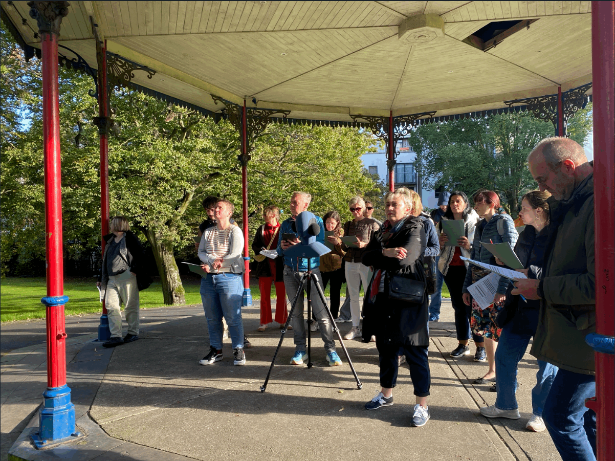 Group of people standing under a park pavilion during an outdoor listening activity.