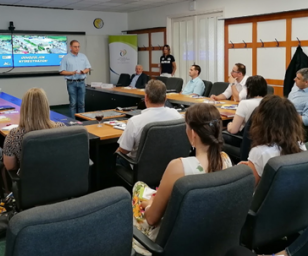 This image shows a meeting or workshop taking place in a conference room. A group of people is seated around a U-shaped table arrangement, facing a presenter who is standing and speaking.