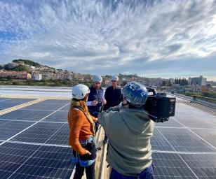 A group of four people on a rooftop covered with solar panels. The setting appears to be an urban area, with buildings and greenery visible in the background under a partly cloudy sky.