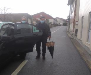 This image shows an elderly man and woman wearing face masks, standing near a taxi parked on a quiet street.