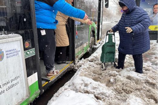 © photo Iris Popescu, 2025. Alt-text: Two people assist an elderly woman with a trolley as she boards a bus, offering their hands to help her navigate a snowy, uncleared bus stop area.