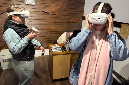 Young test VR headsets branded “URBACT METACITY”; one smiles while holding a controller, the other adjusts the visor.