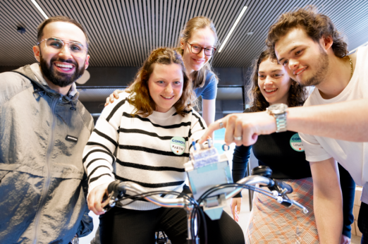 Um grupo de pessoas reunidas em torno de uma bicicleta dentro de casa, concentradas e apontando para um pequeno dispositivo montado no guiador, num ambiente colaborativo.