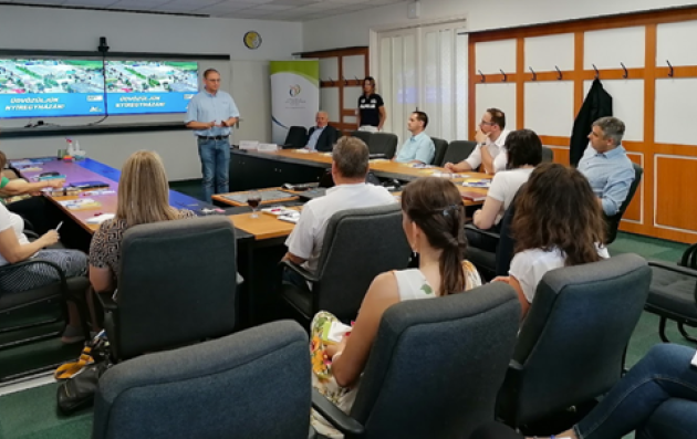 This image shows a meeting or workshop taking place in a conference room. A group of people is seated around a U-shaped table arrangement, facing a presenter who is standing and speaking.