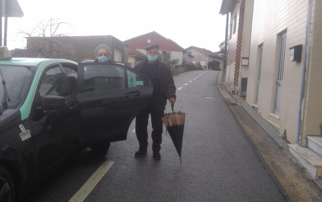 This image shows an elderly man and woman wearing face masks, standing near a taxi parked on a quiet street.