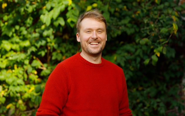 Kalle smiling in red sweater and green, leafy background