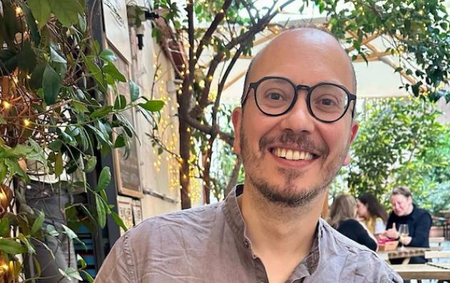 Smiling person, between 40 and 50 years old, with glasses sitting at an outdoor café surrounded by green plants and soft string lights