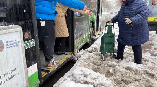 © photo Iris Popescu, 2025. Alt-text: Two people assist an elderly woman with a trolley as she boards a bus, offering their hands to help her navigate a snowy, uncleared bus stop area.