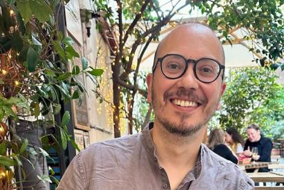 Smiling person, between 40 and 50 years old, with glasses sitting at an outdoor café surrounded by green plants and soft string lights