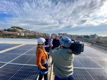 A group of four people on a rooftop covered with solar panels. The setting appears to be an urban area, with buildings and greenery visible in the background under a partly cloudy sky.