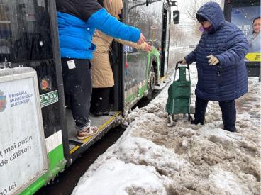© photo Iris Popescu, 2025. Alt-text: Two people assist an elderly woman with a trolley as she boards a bus, offering their hands to help her navigate a snowy, uncleared bus stop area.