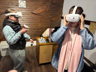 Young test VR headsets branded “URBACT METACITY”; one smiles while holding a controller, the other adjusts the visor.