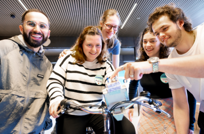 Um grupo de pessoas reunidas em torno de uma bicicleta dentro de casa, concentradas e apontando para um pequeno dispositivo montado no guiador, num ambiente colaborativo.