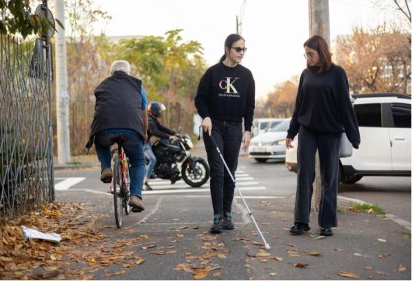 © photo AMAIS, 2022. Alt-text: A visually impaired woman using a white cane walks on a leaf-covered sidewalk, accompanied by another woman. A cyclist and a motorcyclist pass nearby on the street, and cars wait at a crosswalk in the background. The scene highlights shared urban space and the importance of accessibility and safety for all users.